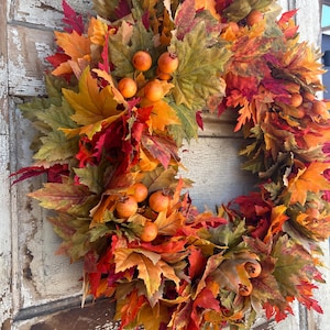 May include: A colourful autumn wreath made from leaves in shades of red, orange, yellow, and green. The wreath is decorated with small, round, orange berries, creating a festive seasonal decoration. The wreath is displayed on a weathered, white-painted wooden door.