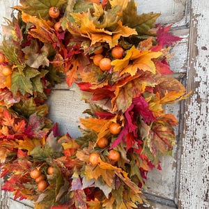 May include: A seasonal wreath featuring autumnal leaves in red, orange, and green hues. The wreath is decorated with small, spherical, orange berries. The leaves are arranged in a circular design, creating a decorative piece for the home.