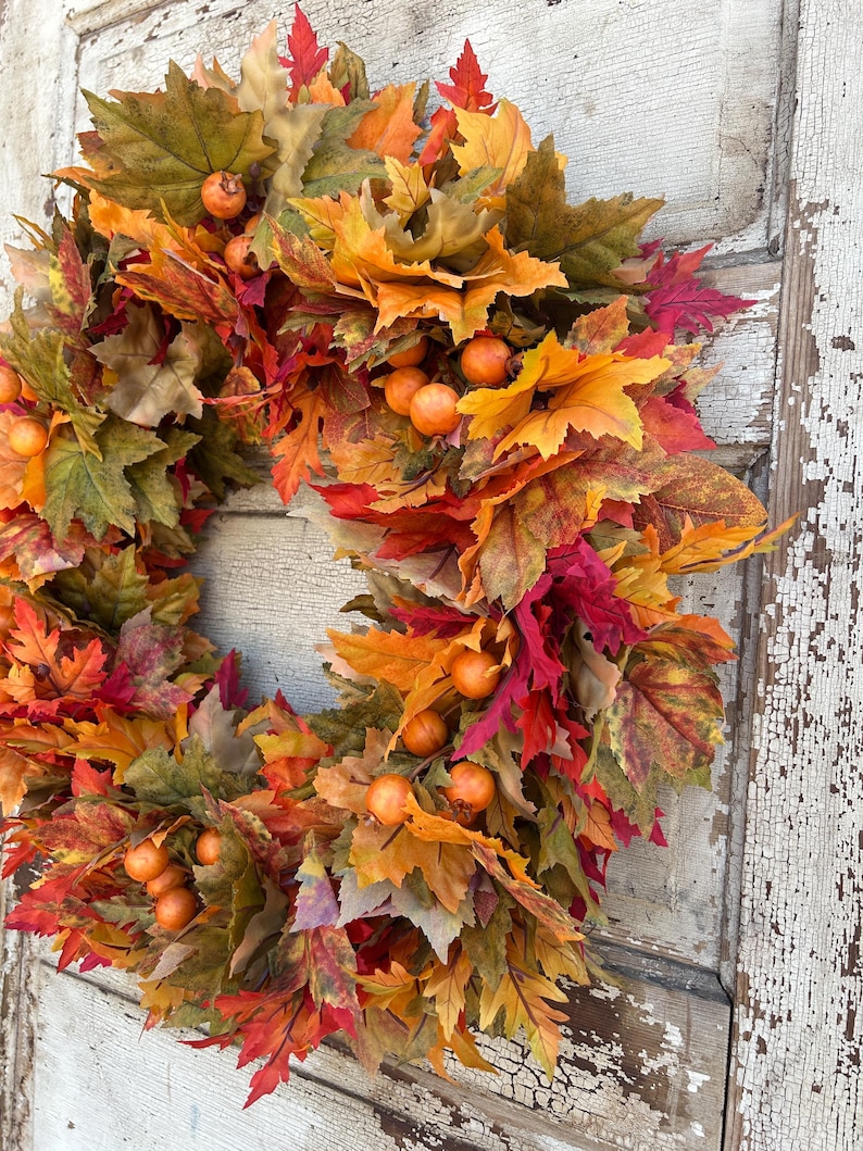 May include: A seasonal wreath featuring autumnal leaves in red, orange, and green hues. The wreath is decorated with small, spherical, orange berries. The leaves are arranged in a circular design, creating a decorative piece for the home.
