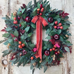 May include: A decorative wreath featuring a mix of greenery, red apples, berries, and a red ribbon. The wreath is composed of various green leaves, with accents of red and burgundy foliage. The apples and berries add pops of color.