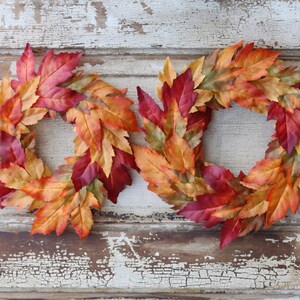 May include: Two autumn leaf wreaths. Each wreath is made of colourful leaves in shades of red, orange, and yellow. The wreaths are arranged side by side against a distressed white and brown wooden door.