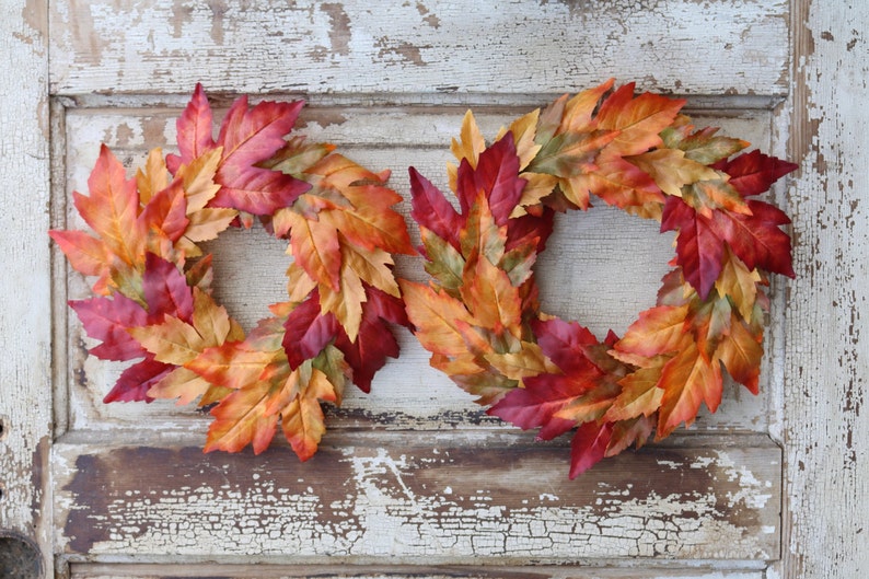 May include: Two autumn leaf wreaths. Each wreath is made of colourful leaves in shades of red, orange, and yellow. The wreaths are arranged side by side against a distressed white and brown wooden door.