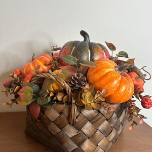 May include: A woven basket filled with decorative pumpkins, gourds, and fall foliage. The pumpkins are various sizes and colors, including orange, green, and yellow. The basket is brown and has a handle.
