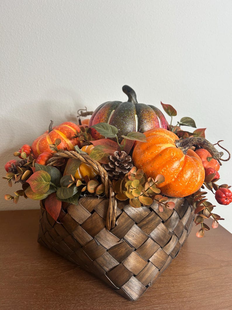 May include: A woven basket filled with decorative pumpkins, gourds, and fall foliage. The pumpkins are various sizes and colors, including orange, green, and yellow. The basket is brown and has a handle.