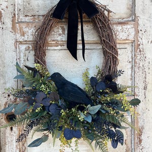 May include: An oval grapevine wreath featuring a black velvet bow at the top and a black crow figurine nestled among dark foliage. The arrangement includes dark blue-black leaves, fern fronds, and small berries, creating a layered, textured appearance.