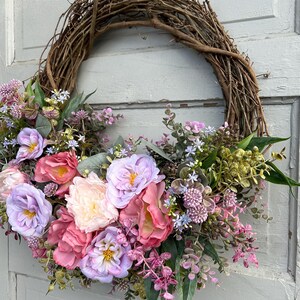 May include: A handcrafted grapevine wreath adorned with an array of faux flowers in shades of pink, lavender, and cream. The floral arrangement includes roses and other blooms, accented by green foliage. The wreath is displayed on a light gray door.