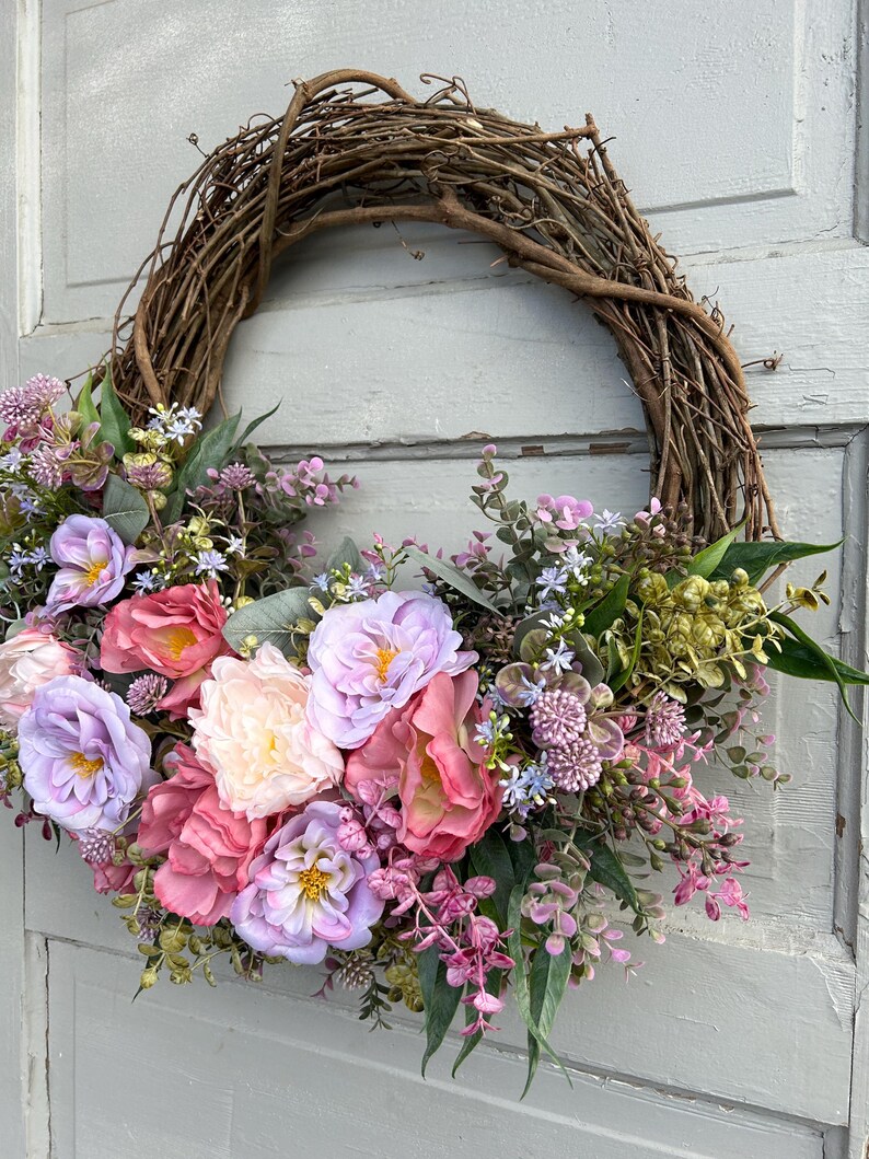 May include: A handcrafted grapevine wreath adorned with an array of faux flowers in shades of pink, lavender, and cream. The floral arrangement includes roses and other blooms, accented by green foliage. The wreath is displayed on a light gray door.