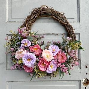 May include: A handcrafted floral wreath featuring a natural grapevine base. The wreath is adorned with an array of colorful faux flowers in shades of pink, purple, and cream, complemented by green foliage. This decorative wreath is perfect for adding a touch of spring to any home.