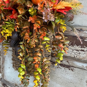 May include: A fall-themed hanging basket overflowing with colorful faux foliage, pumpkins, and mushrooms. The arrangement features a variety of textures and hues, including orange, red, yellow, and green, creating a warm and inviting autumn display.