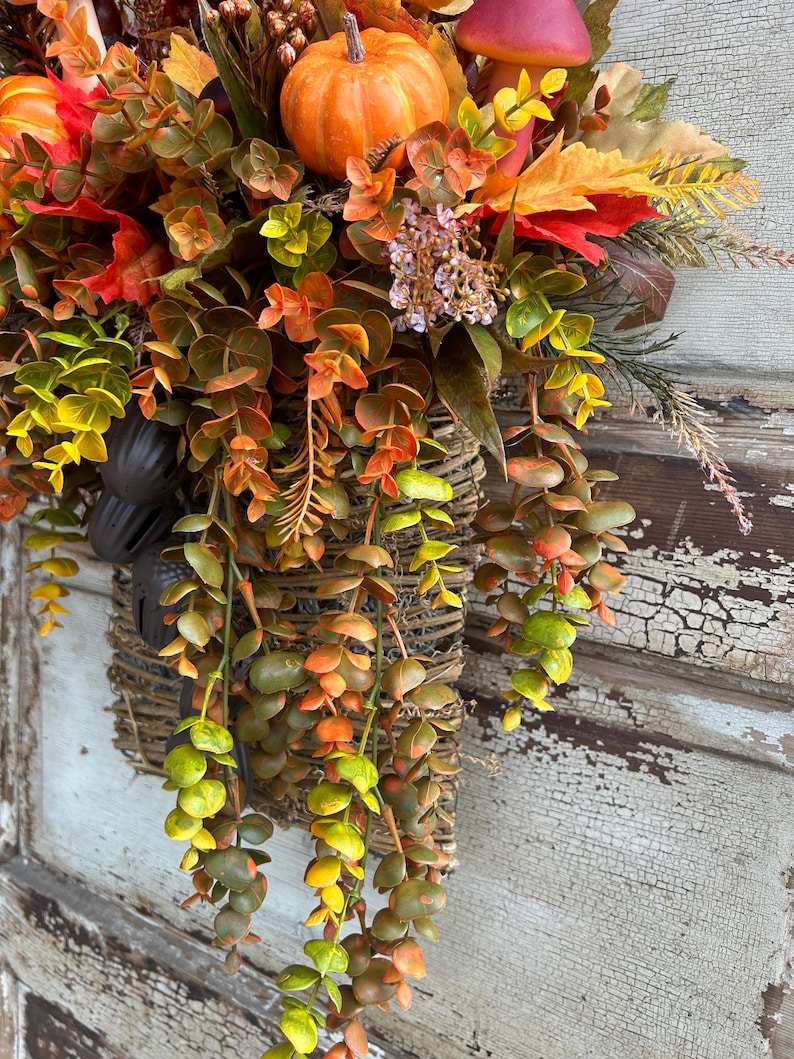 May include: A fall-themed hanging basket overflowing with colorful faux foliage, pumpkins, and mushrooms. The arrangement features a variety of textures and hues, including orange, red, yellow, and green, creating a warm and inviting autumn display.