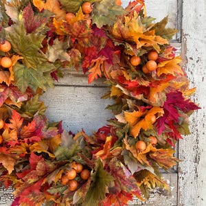 May include: A circular autumn wreath featuring a variety of leaves in autumnal hues of red, orange, yellow, and green. Small, orange berries are dotted amongst the leaves. Ideal for seasonal home decoration.