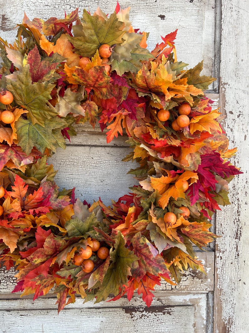 May include: A circular autumn wreath featuring a variety of leaves in autumnal hues of red, orange, yellow, and green. Small, orange berries are dotted amongst the leaves. Ideal for seasonal home decoration.