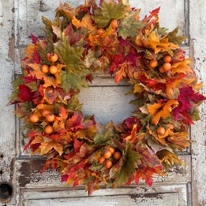 May include: A seasonal wreath featuring autumnal maple leaves in red, orange, and green, with small orange berries. The wreath is set against a backdrop of a distressed, white-painted wooden door.