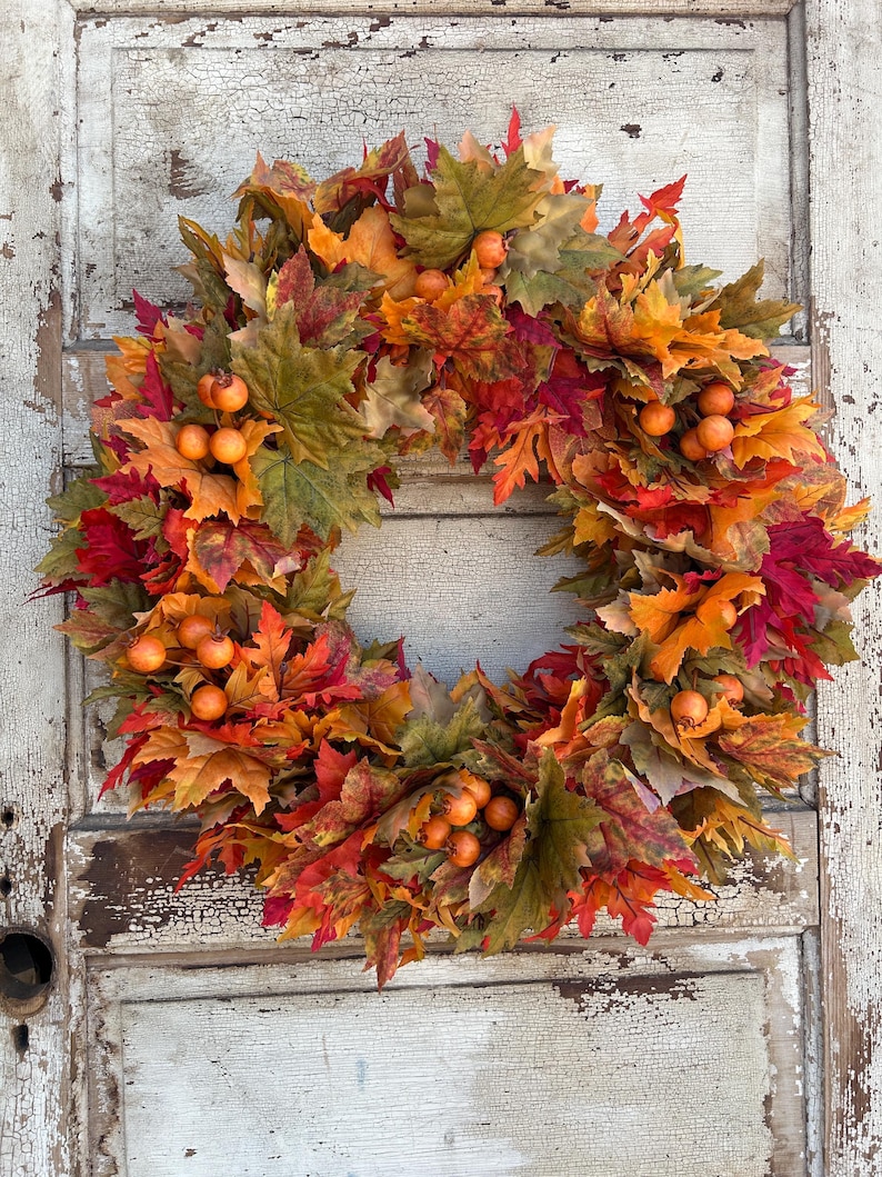 May include: A seasonal wreath featuring autumnal maple leaves in red, orange, and green, with small orange berries. The wreath is set against a backdrop of a distressed, white-painted wooden door.