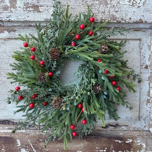 May include: A Christmas wreath featuring green foliage, red berries, and brown pine cones. The wreath is displayed on a distressed, white-painted wooden door, creating a rustic festive decoration.