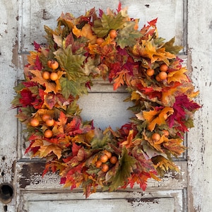 May include: A fall-themed wreath composed of colorful faux maple leaves in shades of red, orange, and green, accented with small orange berries. The wreath is displayed on a distressed, white-painted wooden door, creating a rustic aesthetic.