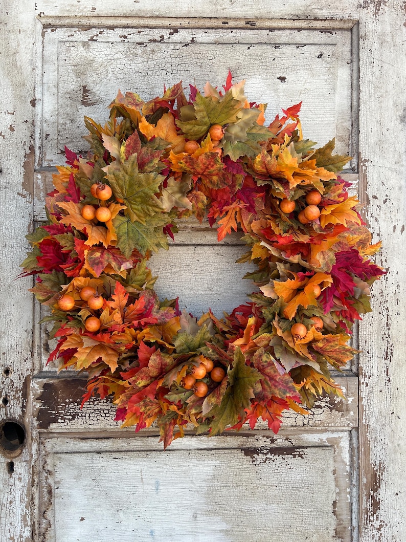 May include: A vibrant autumn wreath featuring artificial maple leaves in shades of red, orange, and green, with small orange berries. The wreath is displayed on a distressed, white-painted wooden door, creating a rustic, autumnal display.