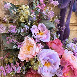 May include: A close-up of a floral wreath with a variety of flowers in shades of pink, purple, and peach. The wreath has a woven base and a purple velvet ribbon. The flowers include peonies and other blooms.