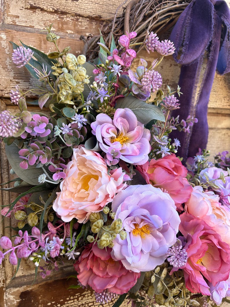 May include: A close-up of a floral wreath with a variety of flowers in shades of pink, purple, and peach. The wreath has a woven base and a purple velvet ribbon. The flowers include peonies and other blooms.
