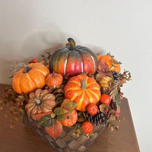 May include: A decorative autumn arrangement featuring various sizes of orange, red, and green pumpkins. The pumpkins are nestled in a woven basket with fall foliage, pinecones, and berries, creating a festive centerpiece.