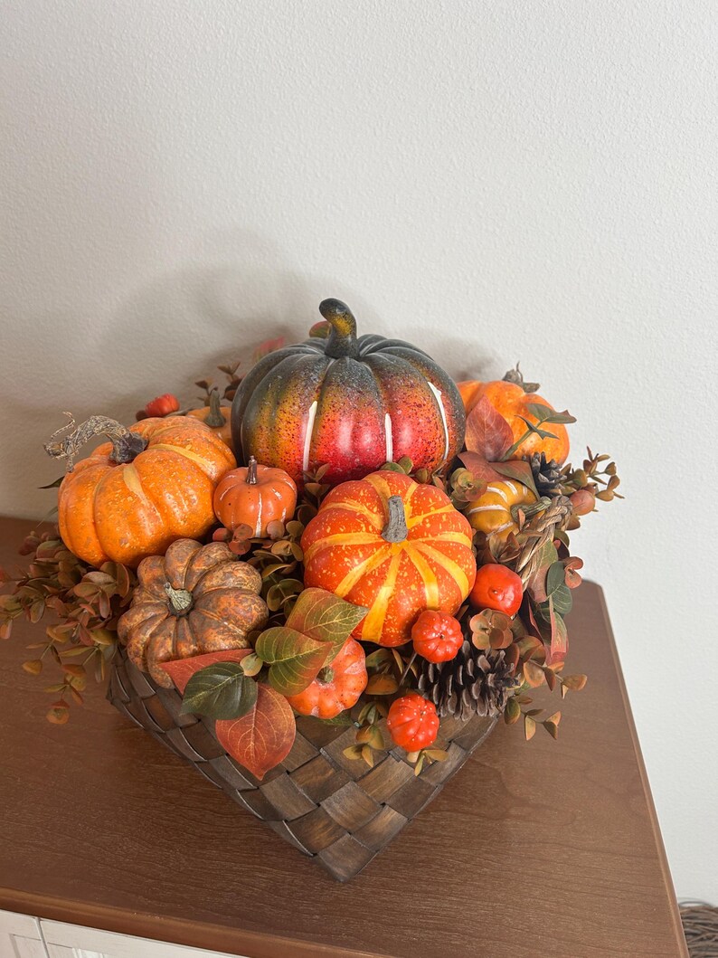 May include: A decorative autumn arrangement featuring various sizes of orange, red, and green pumpkins. The pumpkins are nestled in a woven basket with fall foliage, pinecones, and berries, creating a festive centerpiece.