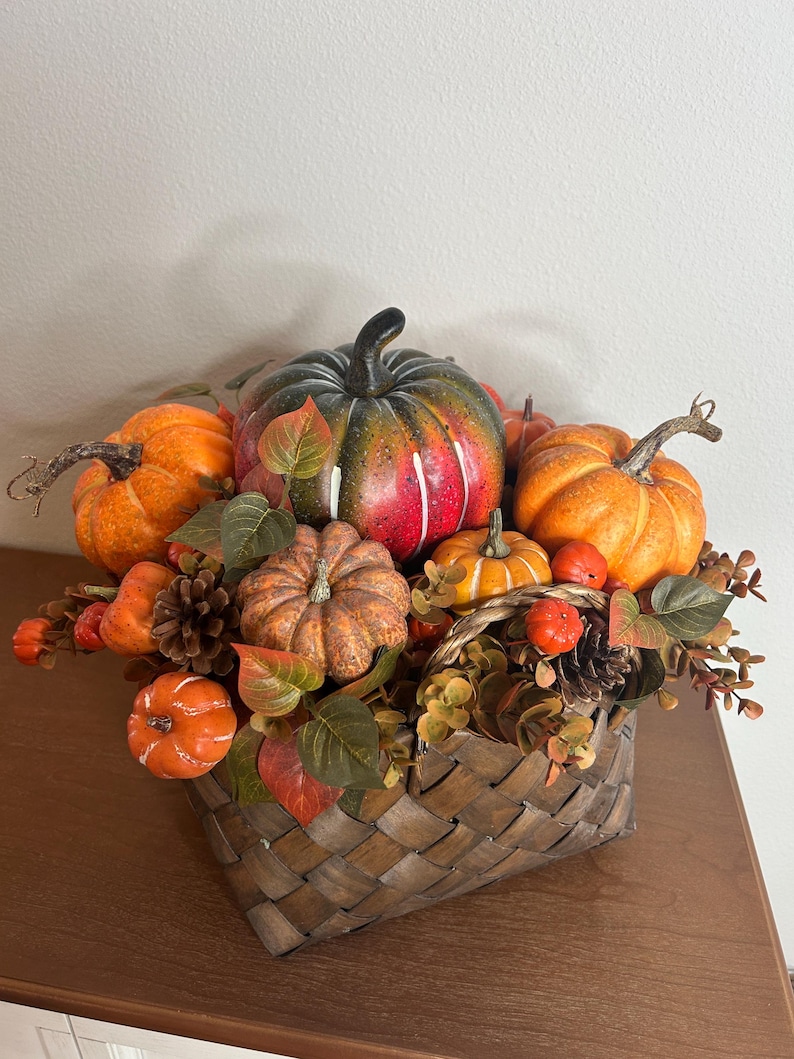 May include: A fall-themed arrangement in a woven basket. The arrangement features various sizes of orange, green, and red pumpkins, along with fall foliage and pinecones. The basket is brown and sits on a wooden surface.
