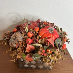 May include: A fall-themed arrangement in a woven basket. The arrangement features vibrant orange pumpkins, red berries, and autumn-colored foliage. The basket is brown and has a woven design, creating a rustic aesthetic.