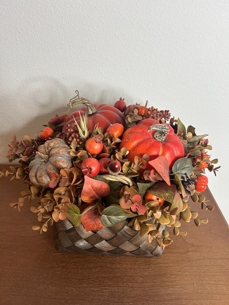 May include: A fall-themed arrangement in a woven basket. The arrangement features vibrant orange pumpkins, red berries, and autumn-colored foliage. The basket is brown and has a woven design, creating a rustic aesthetic.