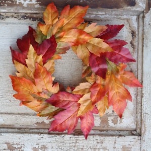 May include: A circular wreath made of artificial autumn leaves in red, orange, and yellow hues. The leaves are arranged to create a decorative piece. The wreath is set against a distressed white wooden background, adding to its rustic charm.