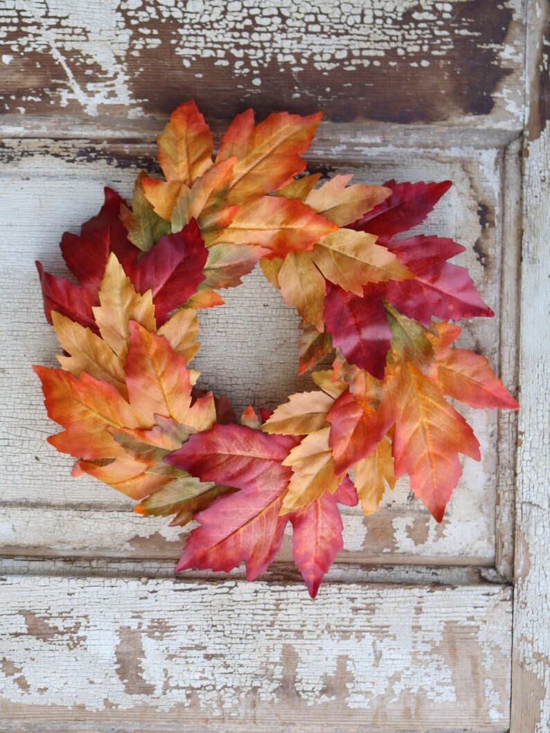 May include: A circular wreath made of artificial autumn leaves in red, orange, and yellow hues. The leaves are arranged to create a decorative piece. The wreath is set against a distressed white wooden background, adding to its rustic charm.