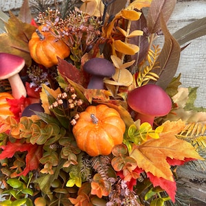 May include: A fall-themed arrangement featuring miniature orange pumpkins, burgundy mushrooms, and colorful autumn leaves. The arrangement includes various foliage in shades of green, yellow, and red, creating a vibrant and seasonal display. The overall theme is a harvest or Thanksgiving decoration.