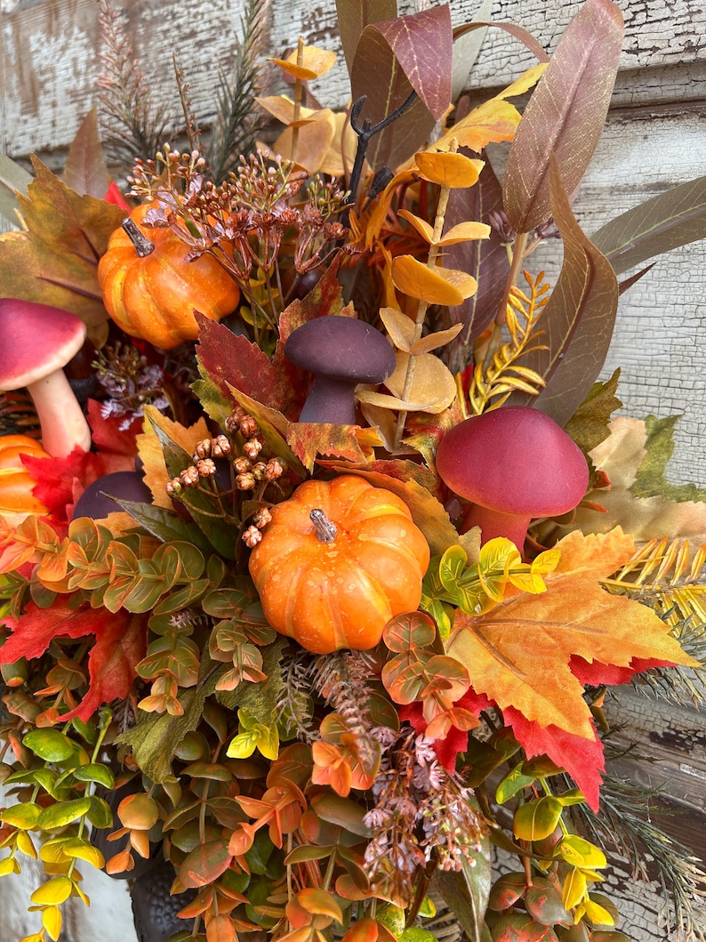 May include: A fall-themed arrangement featuring miniature orange pumpkins, burgundy mushrooms, and colorful autumn leaves. The arrangement includes various foliage in shades of green, yellow, and red, creating a vibrant and seasonal display. The overall theme is a harvest or Thanksgiving decoration.
