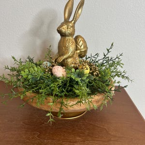 May include: A gold-colored rabbit statue with an egg, nestled in a wooden bowl filled with greenery and decorative eggs. The Easter-themed centerpiece is set on a wooden surface.