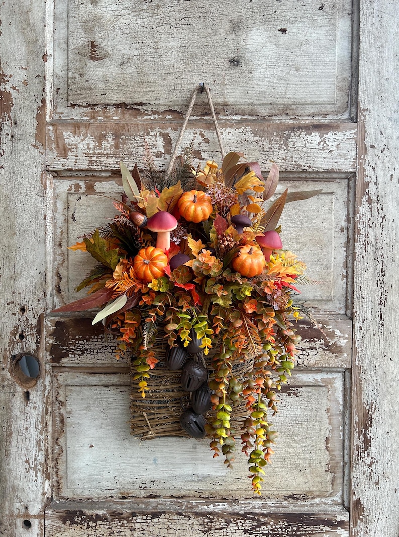 May include: A fall-themed wall hanging featuring a woven basket filled with pumpkins, mushrooms, and autumn foliage. The arrangement includes orange pumpkins, red-capped mushrooms, and various leaves in shades of orange, green, and brown. A jute rope is used for hanging.
