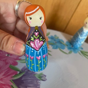 May include: A hand-painted wooden doll with red hair, a white headband, and a blue dress with pink and green floral details.