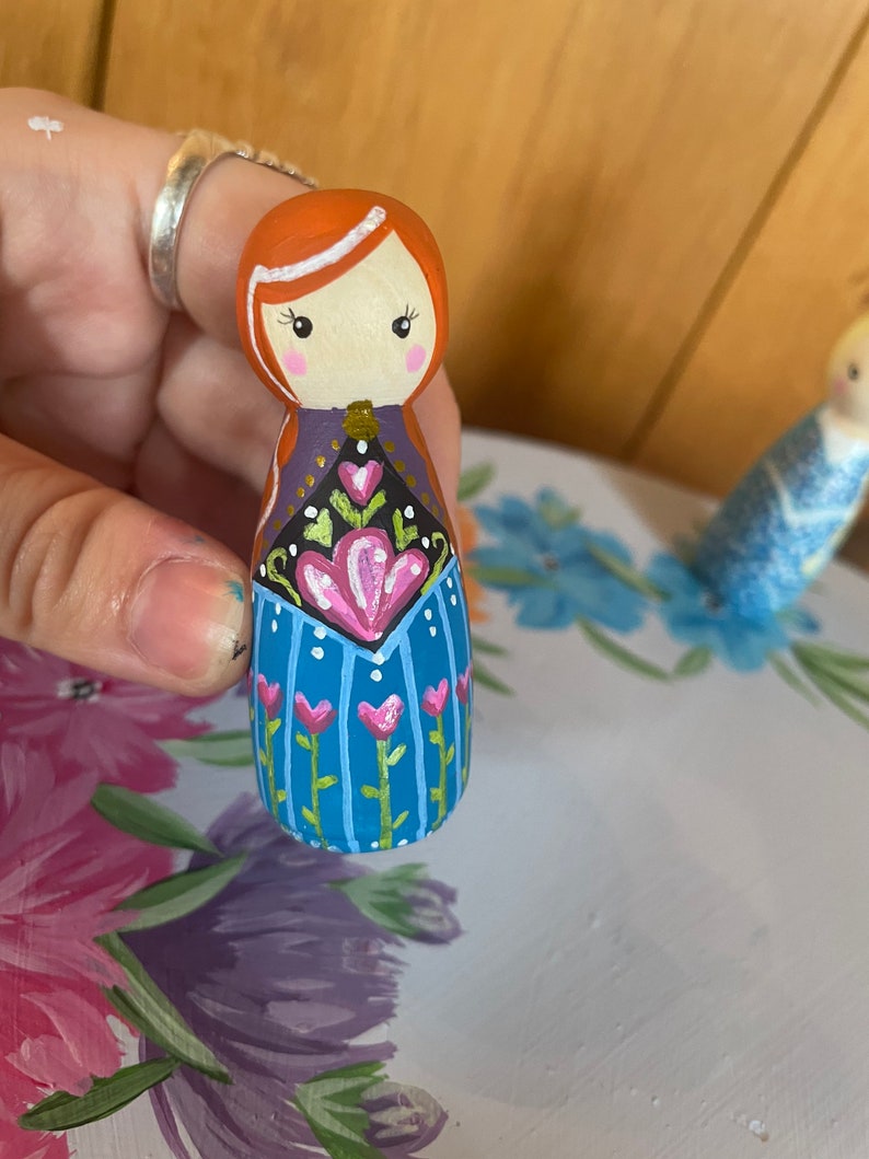 May include: A hand-painted wooden doll with red hair, a white headband, and a blue dress with pink and green floral details.