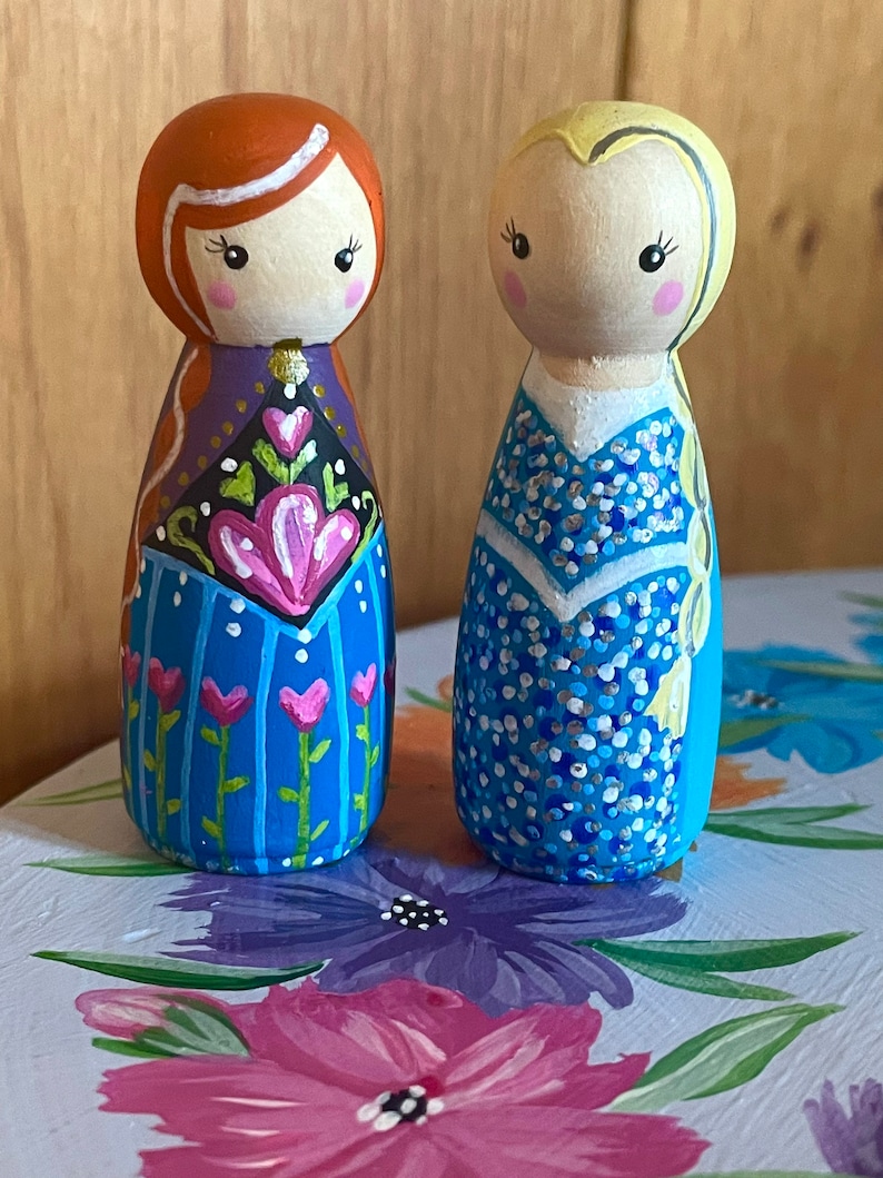 May include: Two hand-painted wooden peg dolls dressed as princesses. The doll on the left has red hair and a blue and purple dress with pink flowers. The doll on the right has blonde hair and a blue dress with white dots.