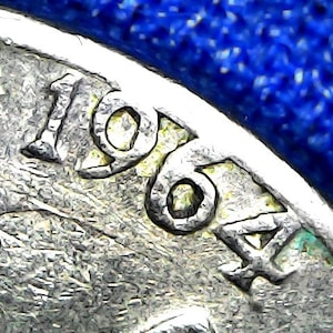 May include: Close-up of a silver coin with the date 1964 visible. The coin's surface has a textured appearance, with the numbers and letters slightly raised. The background is a deep blue color.