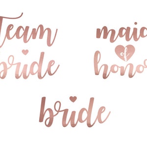Op de afbeelding: Drie ontwerpen in rosé gouden folie voor een bruiloftsfeest. De ontwerpen luiden "Team Bride", "Bride" en "Maid of Honor".