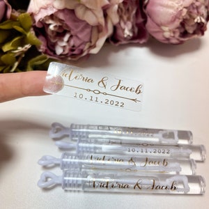 Peut inclure: Plusieurs flacons de bulles transparents avec des poignées en forme de cœur, portant l'inscription dorée "Victoria & Jacob" et la date "10.11.2022". Un autocollant transparent avec le même texte est tenu. Des fleurs roses en arrière-plan.