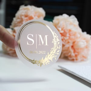 Puede incluir: Pegatina redonda transparente con un diseño de lámina dorada. El diseño presenta una corona de hojas y un círculo con las iniciales "S|M" y la fecha "06.05.2022".