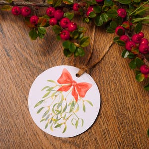 Könnte beinhalten: Ein rundes, weißes Geschenkanhänger-Schild mit einer Aquarellillustration von grünen Mistelzweigen, die mit einer leuchtend roten Schleife gebunden sind. Der Anhänger hat ein Loch zum Aufhängen und ist an einer dünnen braunen Schnur befestigt.