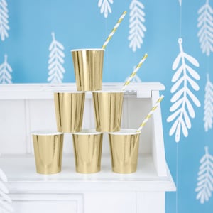 May include: A stack of six gold paper cups with white interiors, arranged on a white shelf. Two cups have gold and white striped straws. The background is a light blue wall with white leaf-shaped decorations.