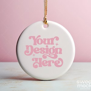May include: White ceramic bauble with a gold string hanging from the top. The bauble has a pink design that says "Your Design Here" in a retro font.