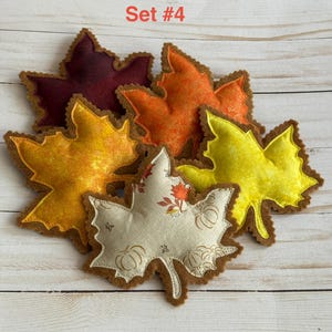 May include: A collection of five fabric maple leaf ornaments labeled Set #4. The leaves feature various colors including deep burgundy, bright orange, golden yellow, and a cream-colored leaf with a brown pumpkin and leaf print pattern.