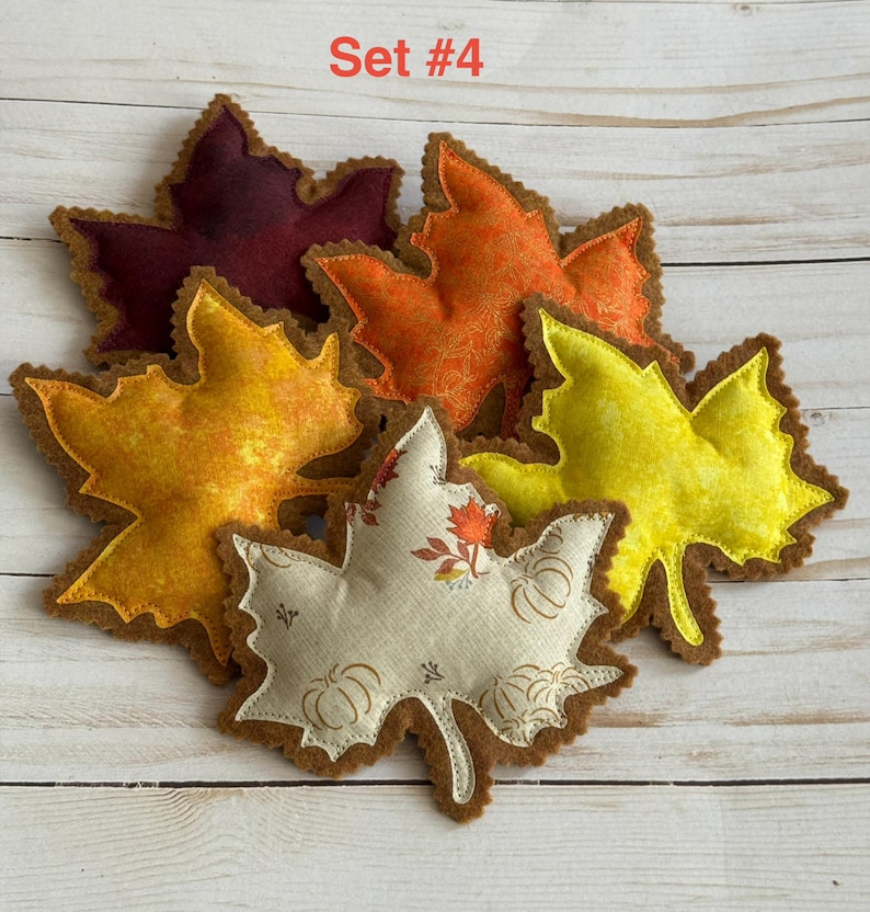 May include: A collection of five fabric maple leaf ornaments labeled Set #4. The leaves feature various colors including deep burgundy, bright orange, golden yellow, and a cream-colored leaf with a brown pumpkin and leaf print pattern.