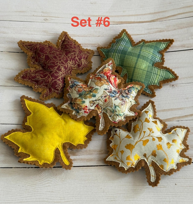 May include: A set of five fabric leaf ornaments labeled Set #6. The collection includes a solid yellow leaf, a maroon leaf with gold filigree, a green plaid leaf, a white leaf with a colorful floral pattern, and a white leaf with gold ginkgo prints.