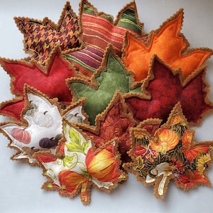 May include: A collection of ten felt maple leaf ornaments in various colors and patterns, including red, green, orange, brown, and white. The leaves are decorated with pumpkins, plaid, and floral designs.