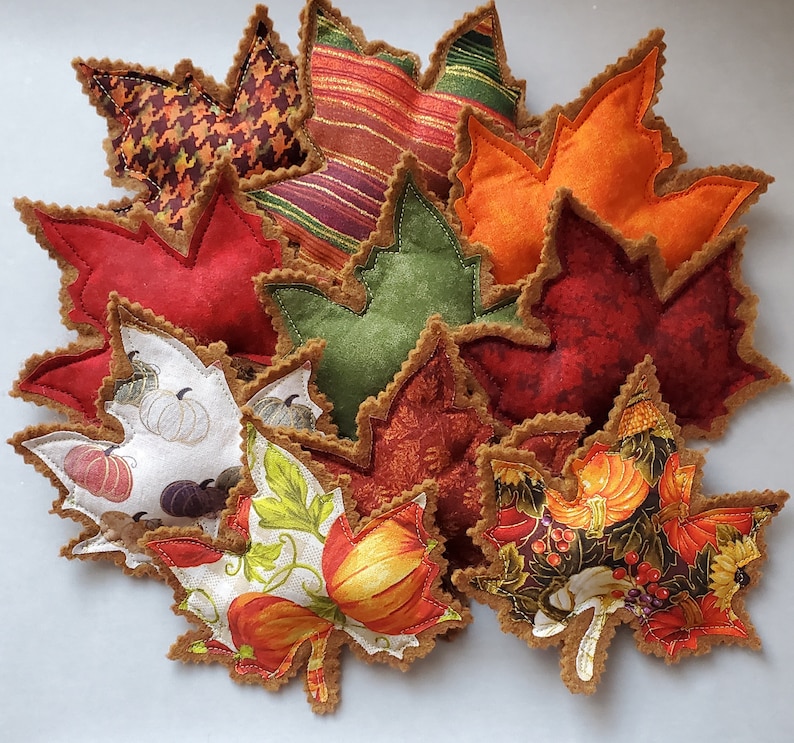 May include: A collection of fabric leaf-shaped ornaments featuring various autumn-themed patterns including pumpkins, houndstooth, stripes, and solid colors in red, orange, green, and brown, all finished with a decorative scalloped felt edge.