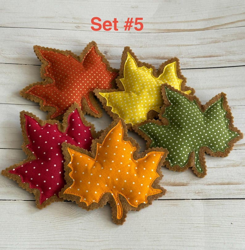 May include: A set of five fabric maple leaf ornaments labeled Set #5. Each leaf features a polka dot pattern in orange, yellow, green, red, and gold, with stitched edges and a soft, stuffed appearance, arranged in a cluster on a light wood surface.